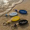 Four SaltBred keychains on a sandy beach with ocean waves.