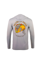 A light gray hooded long-sleeve shirt shown from the back, featuring a large graphic of a manatee inside a taco with the text 'TACOS GO GOOD' and 'I'M ALMOST ILLEGAL'.