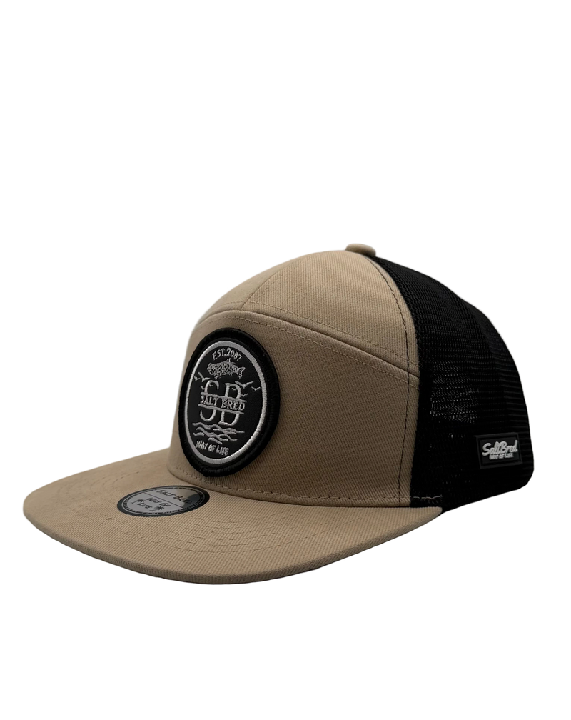 A tan and black trucker hat with a black mesh back, a round black and white embroidered patch on the front, and a small black 'Self-Ad' brand tag on the side.
