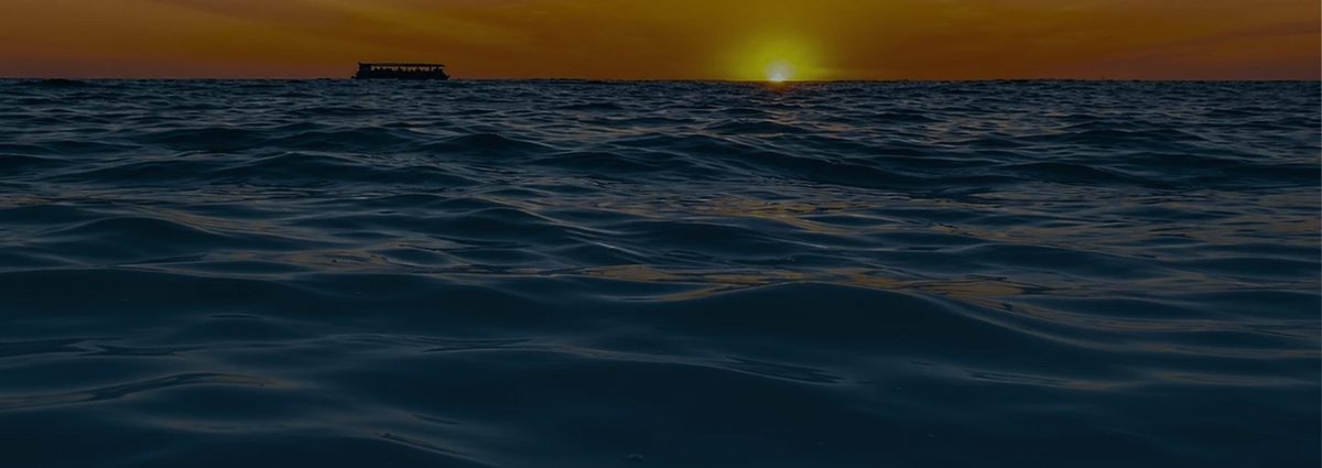 A dark ocean sunset scene with gentle waves in the foreground, a small boat silhouette on the horizon, and the sun low over the water.